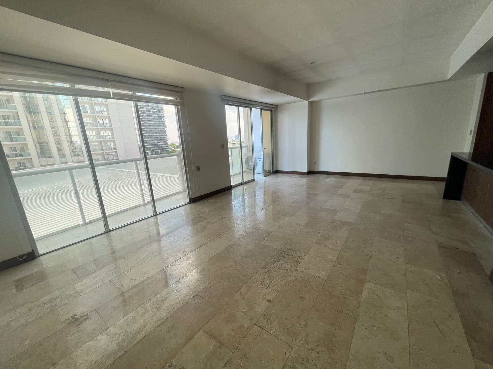 Sala vacía con piso de mármol, paredes blancas y grandes ventanales con vista a edificios.