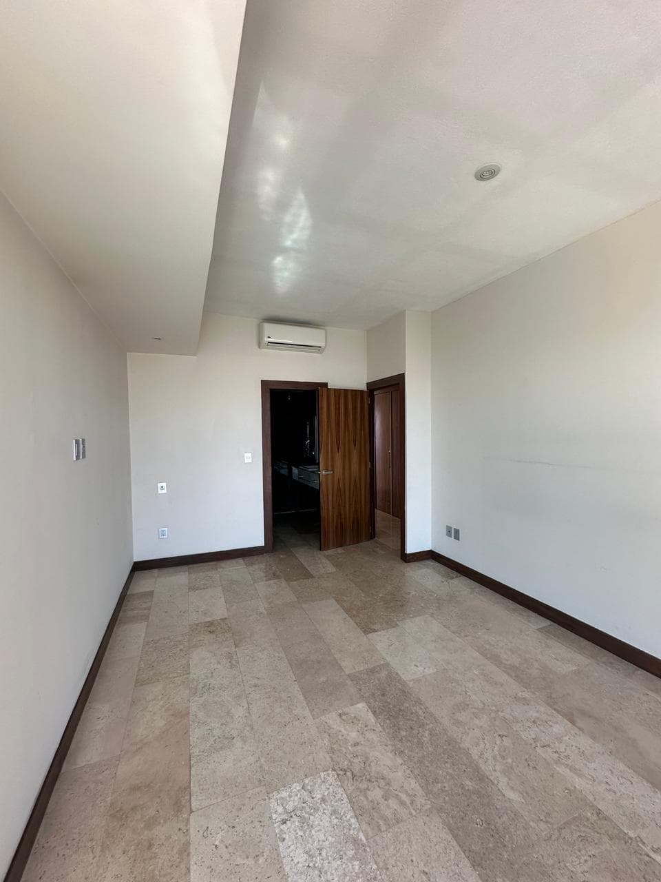 Habitación vacía con piso de baldosas claras, paredes blancas, puerta de madera y aire acondicionado.