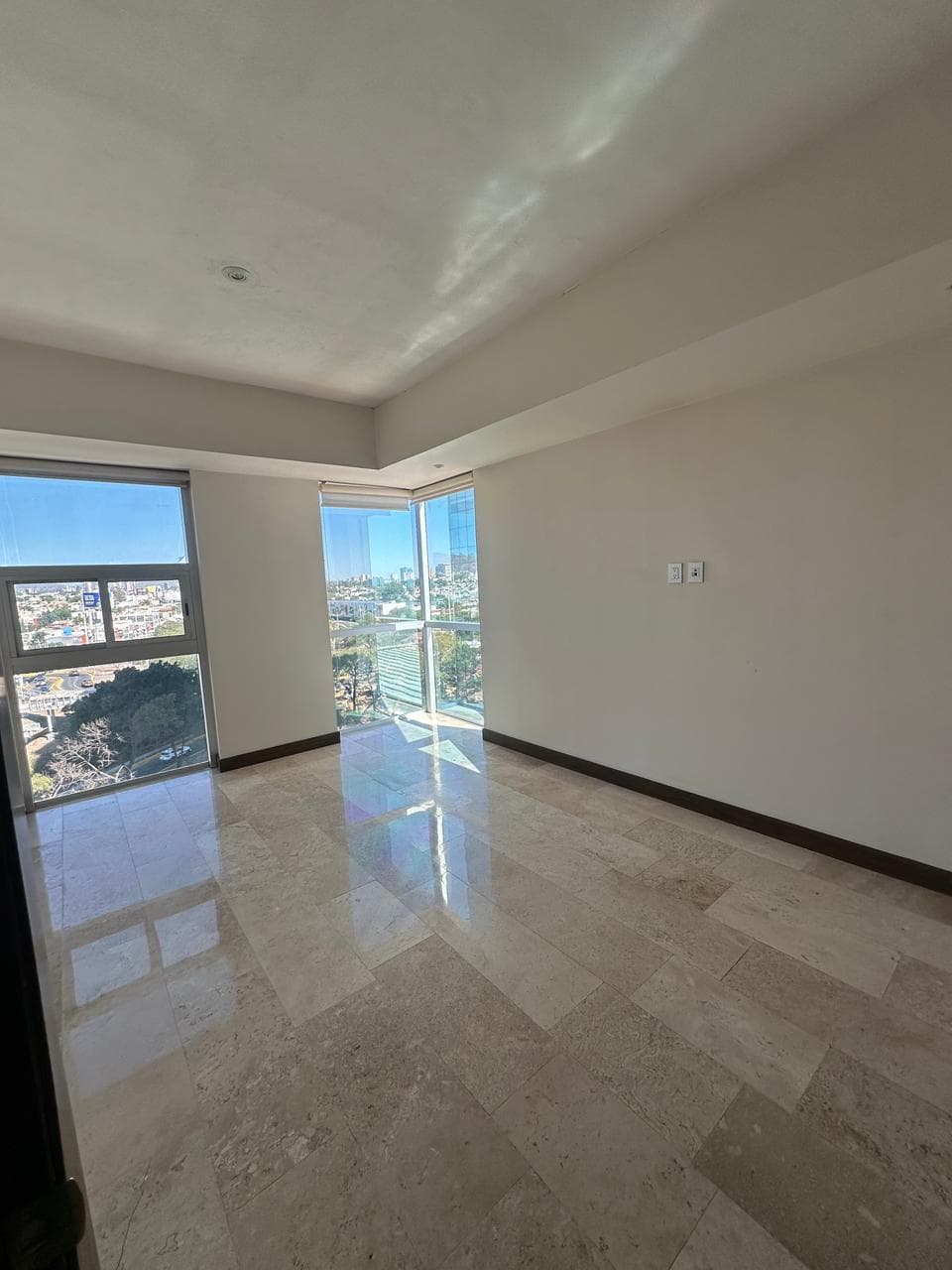 Habitación vacía iluminada con suelos de mármol, paredes blancas y ventanales con vistas urbanas.