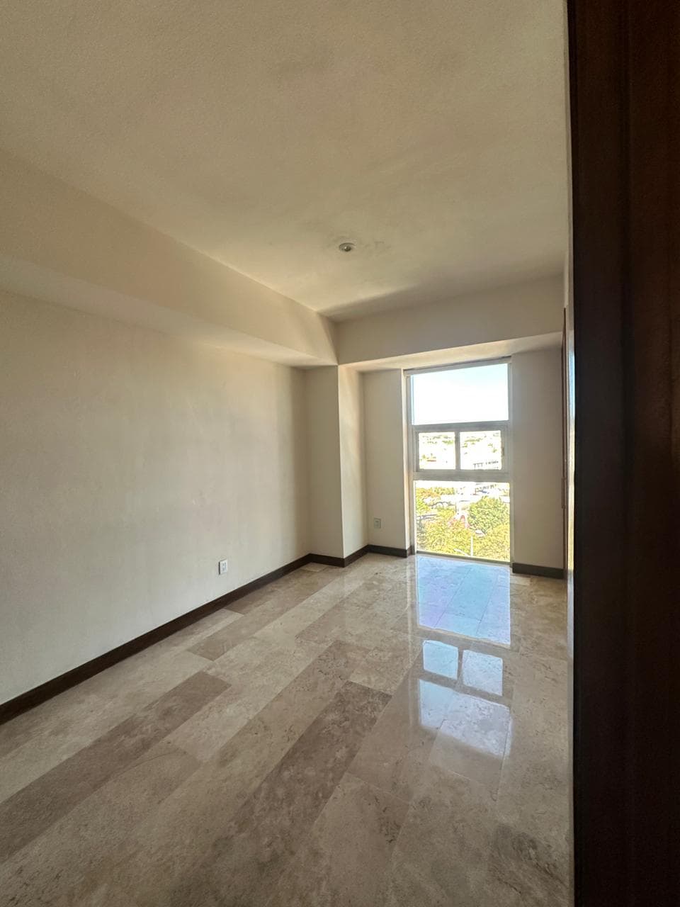 Habitación vacía con paredes color crema, suelo de mármol pulido y una ventana al fondo.
