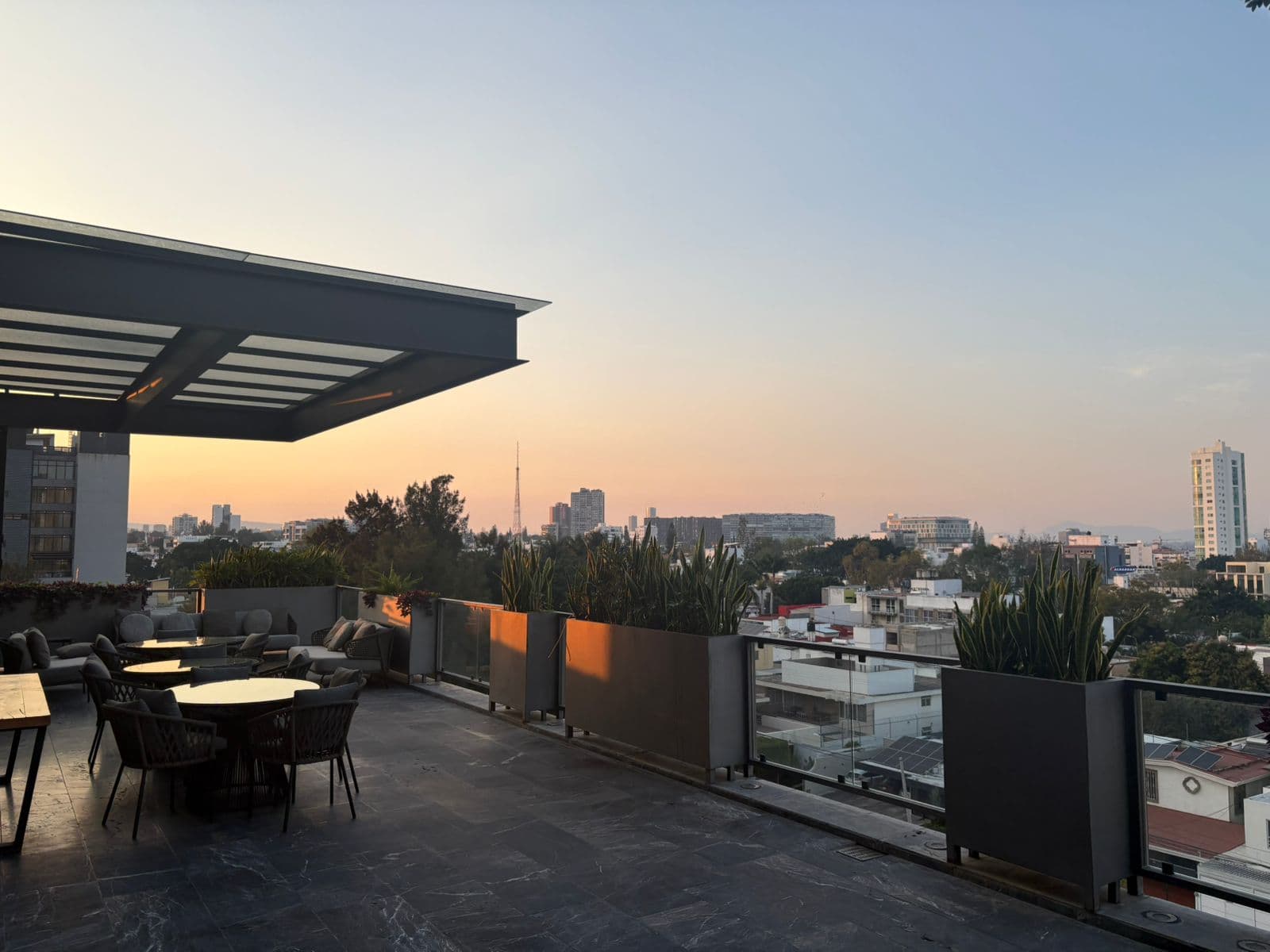 Terraza moderna con mesas y plantas frente a un horizonte urbano al atardecer.