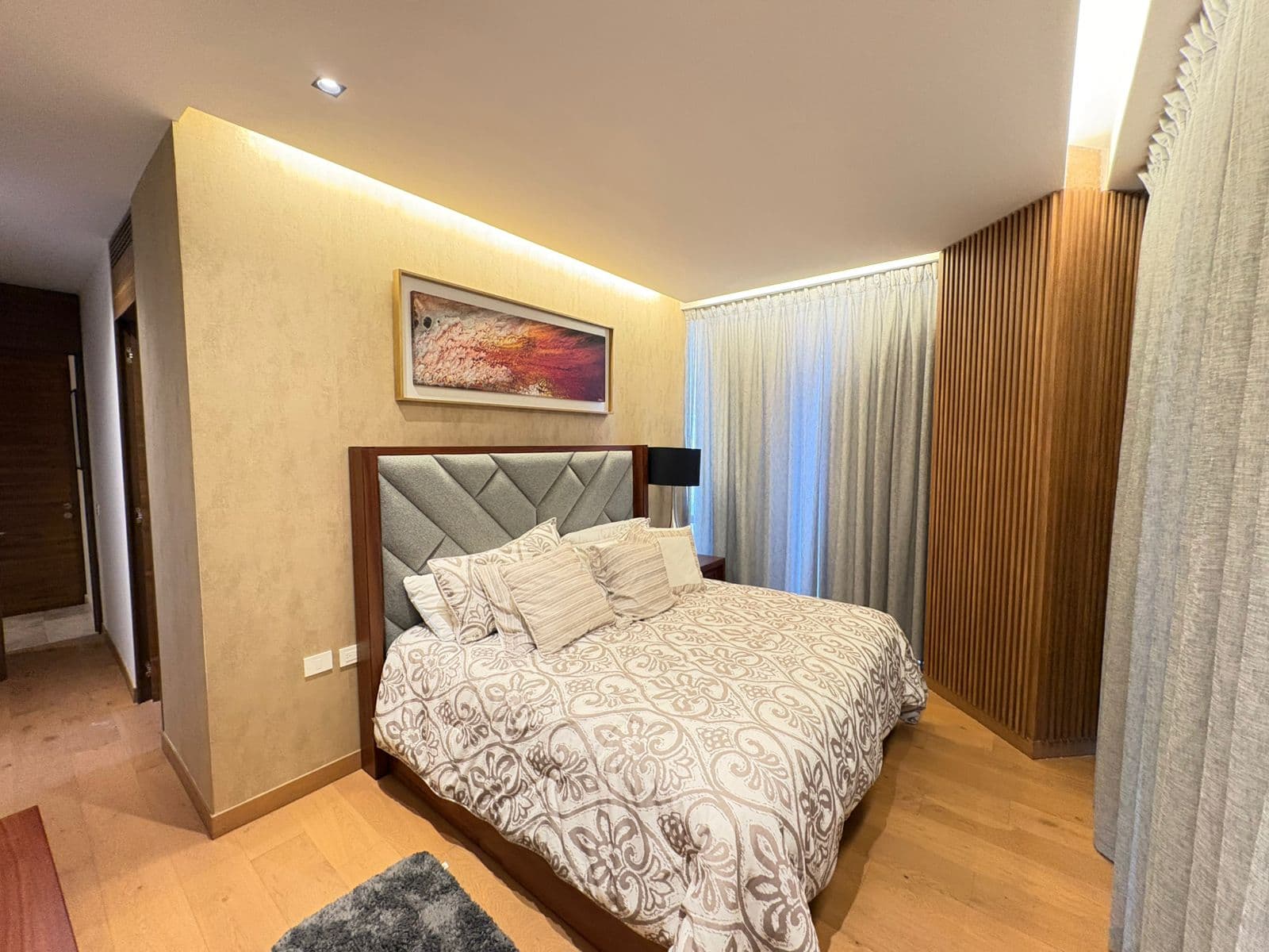 Dormitorio moderno con cama grande, cabecero gris, cuadro abstracto y luz cálida indirecta.