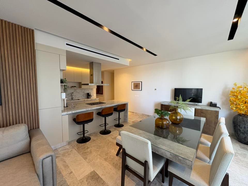Comedor moderno con mesa de cristal, cocina abierta con barra y taburetes de madera elegantes.