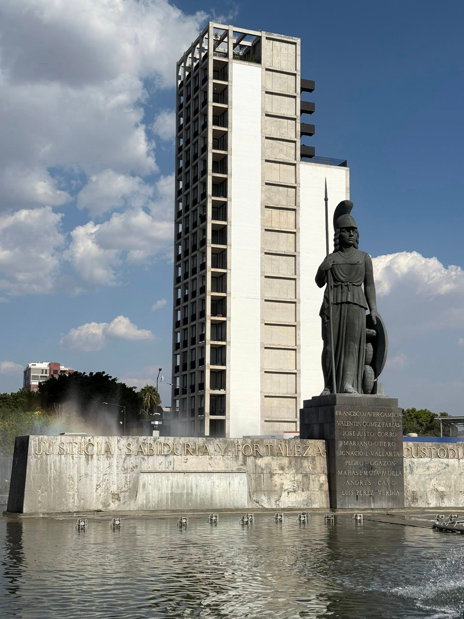 Monumento a la Minerva con lanza y escudo frente a un edificio moderno y fuente.