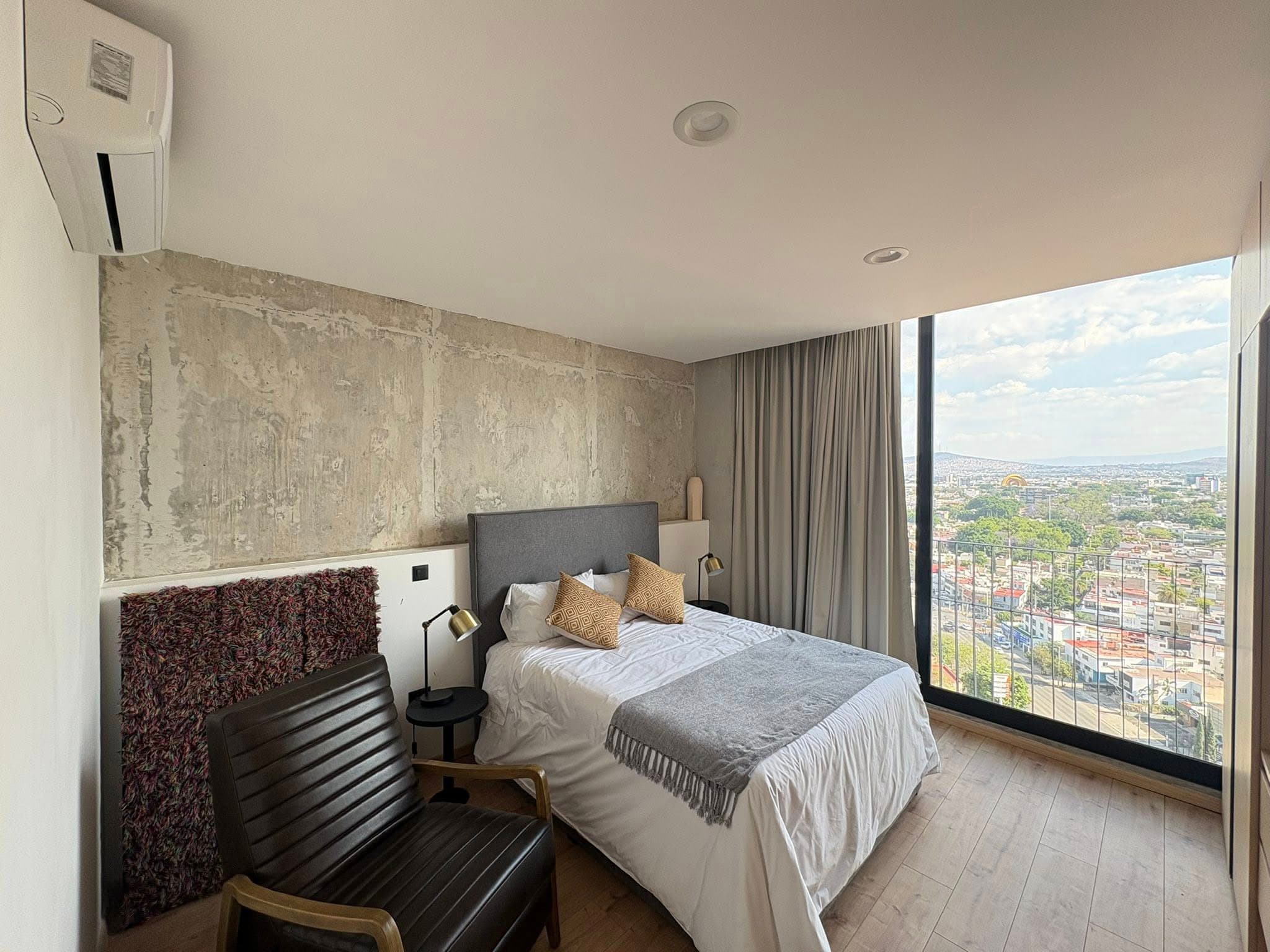 Dormitorio con pared de concreto, cama, sillón negro y ventanal con vistas a la ciudad.