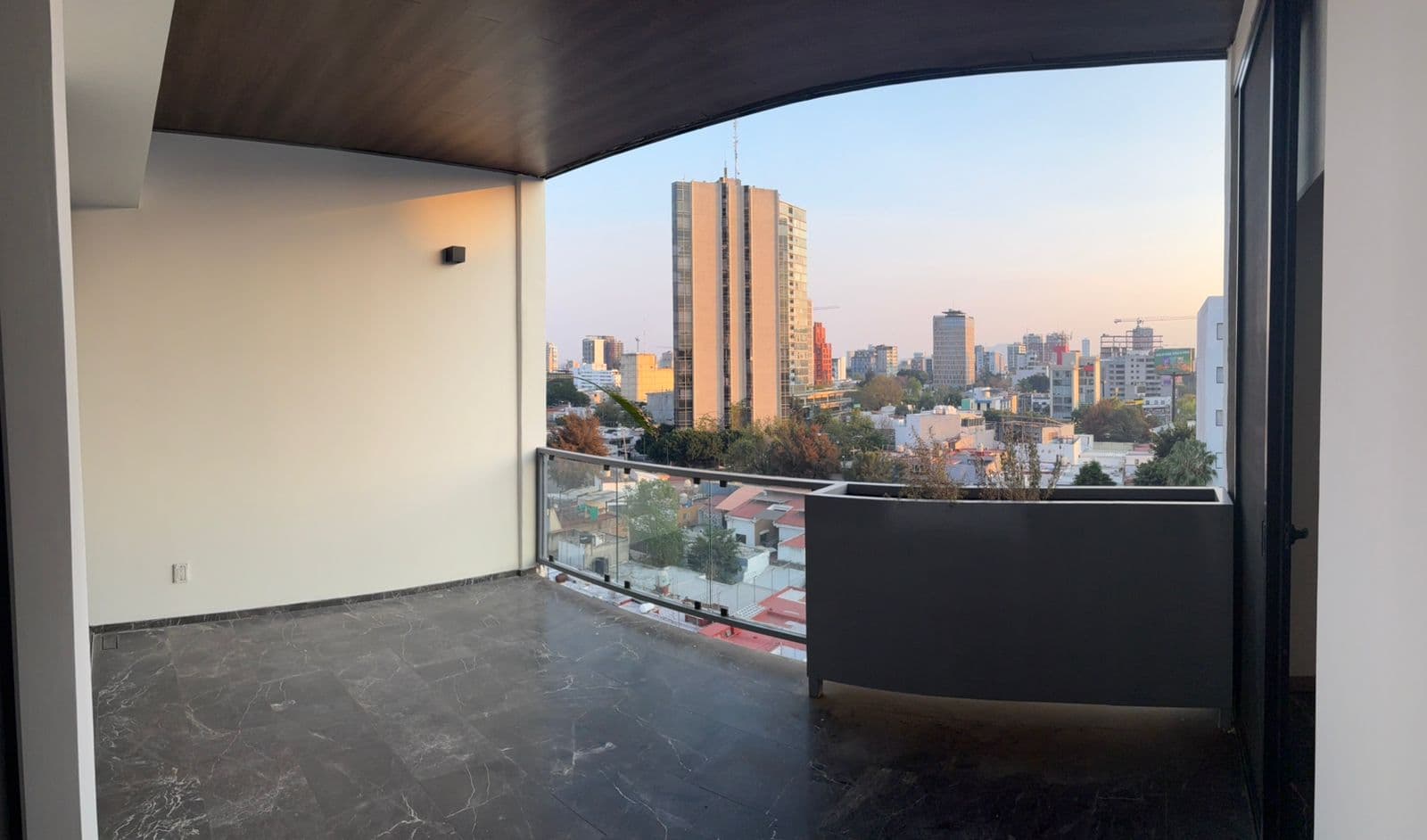 Balcón moderno con suelo oscuro y vista panorámica de edificios urbanos durante el atardecer.