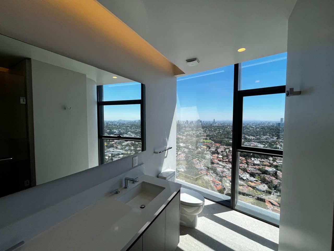 Baño moderno con lavabo, espejo y gran ventanal con vistas panorámicas a la ciudad.