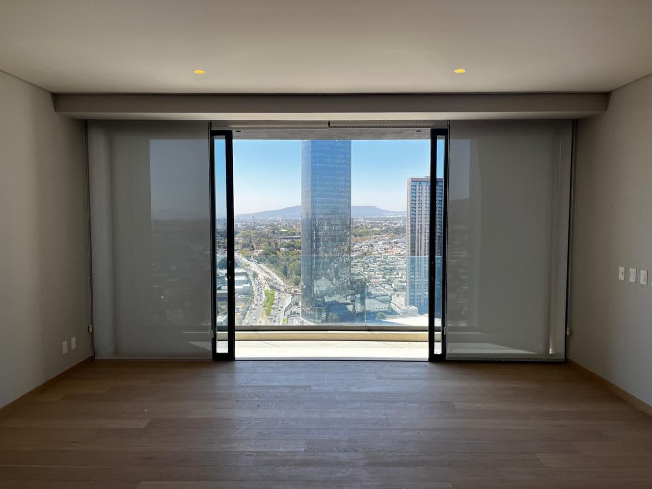 Habitación vacía con suelos de madera y ventanal con vistas a rascacielos y montañas.