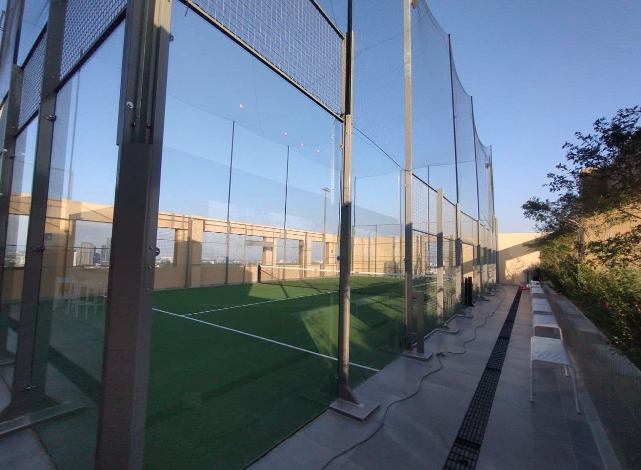 Pista de pádel en azotea con paredes de cristal, césped artificial verde y bancos blancos.